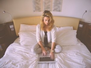 Blonde woman sitting on her bed whilst working on her laptop - flexible working
